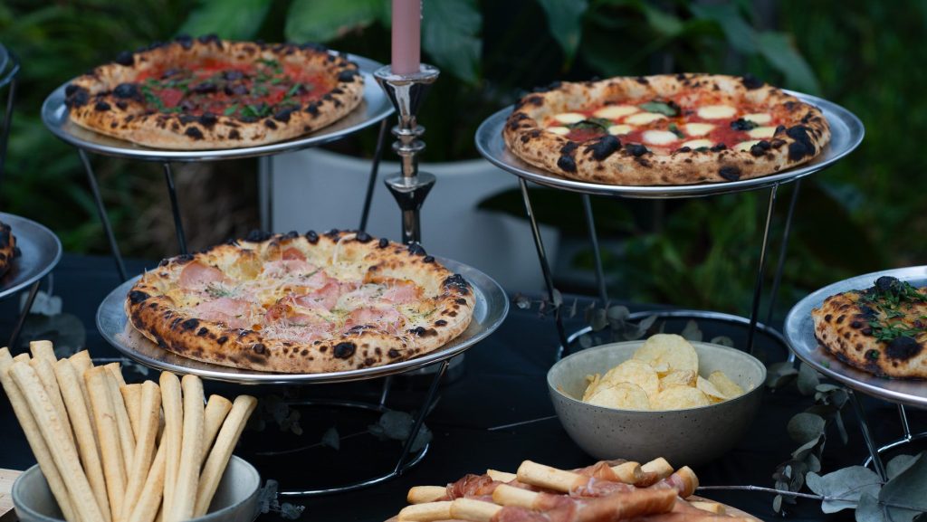 Wood-fired pizza catering spread featuring assorted pizzas, grissini, and prosciutto at an outdoor event in Wollongong.